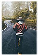 Start Here path visual, man walking down winding road with hands in his pockets, back to us, has on a tan hat and red jacket, black pants.